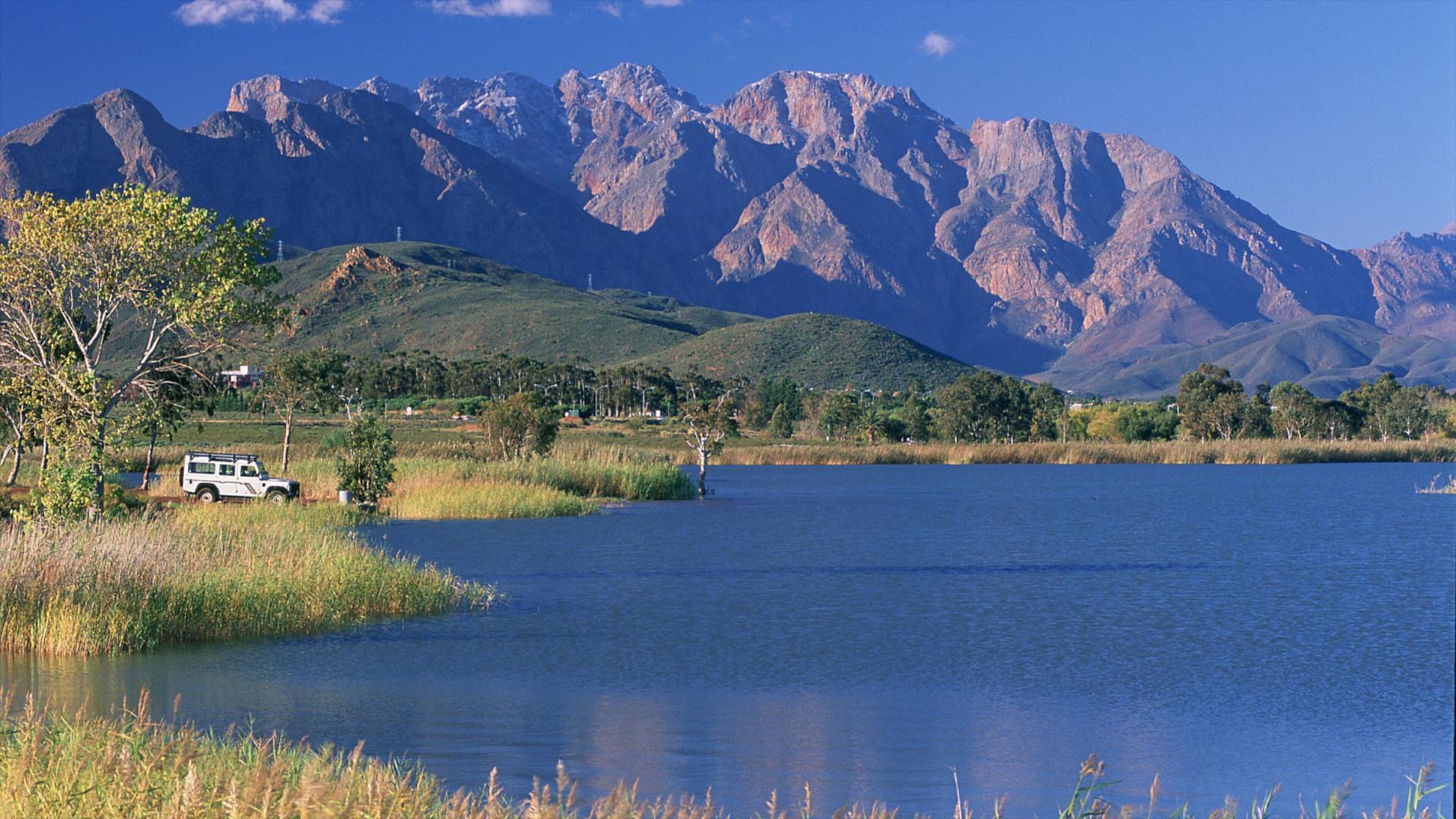 A lake with mountains in the background, a white vehicle parked by the water, and greenery around.