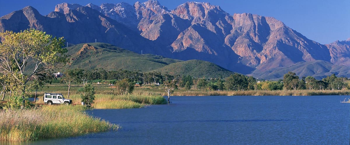 A lake with mountains in the background, a white vehicle parked by the water, and greenery around.