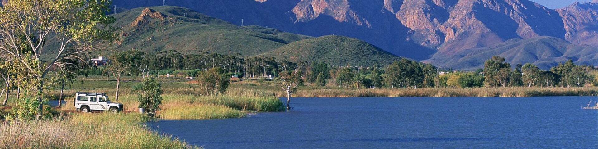 A lake with mountains in the background, a white vehicle parked by the water, and greenery around.