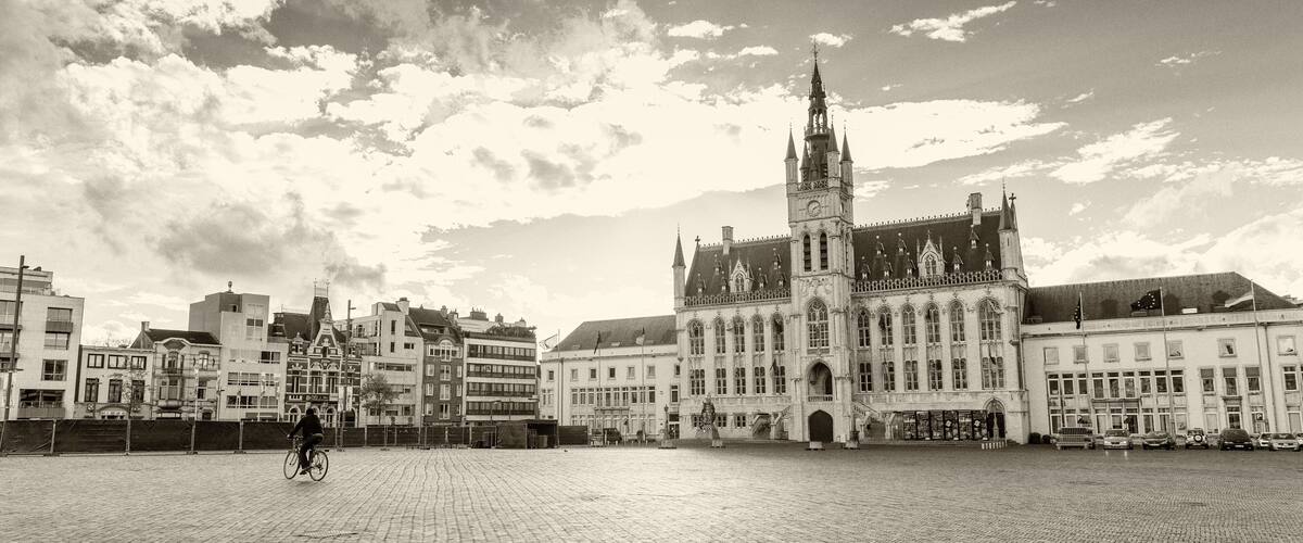 A historic building with a tall tower, a paved square, and modern buildings in the background.