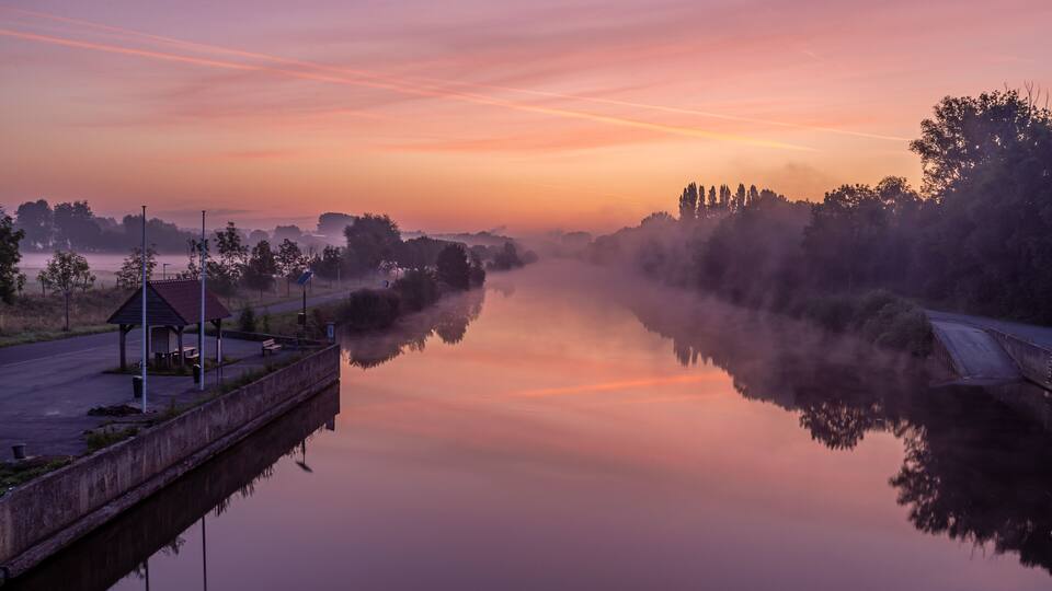 A calm river at dawn with mist, a gazebo, and trees reflecting in the water.