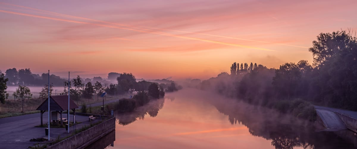 A calm river at dawn with mist, a gazebo, and trees reflecting in the water.