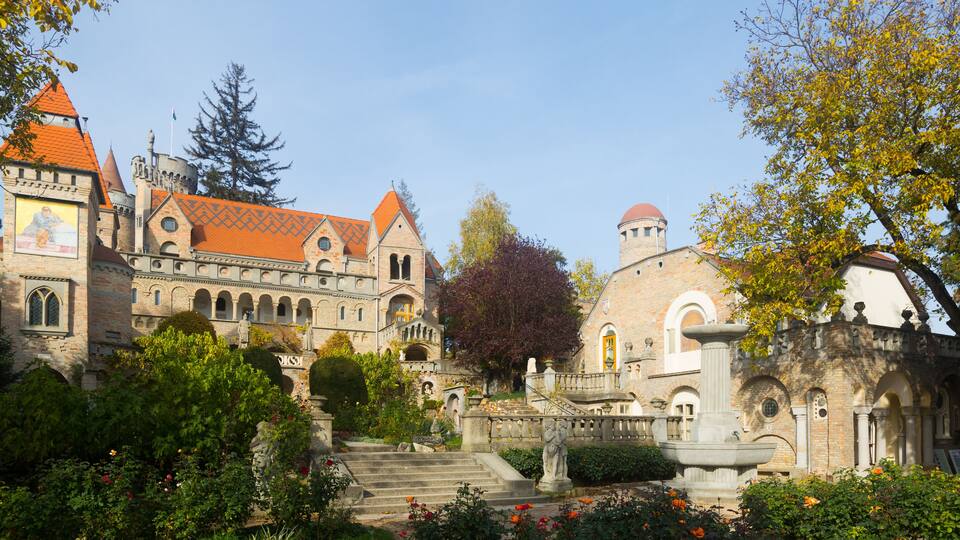 A historic building with a red-tiled roof, a statue, and a fountain in a garden.