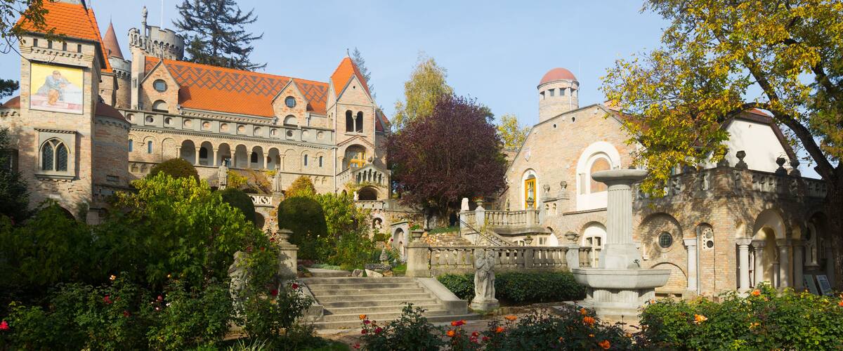 A historic building with a red-tiled roof, a statue, and a fountain in a garden.