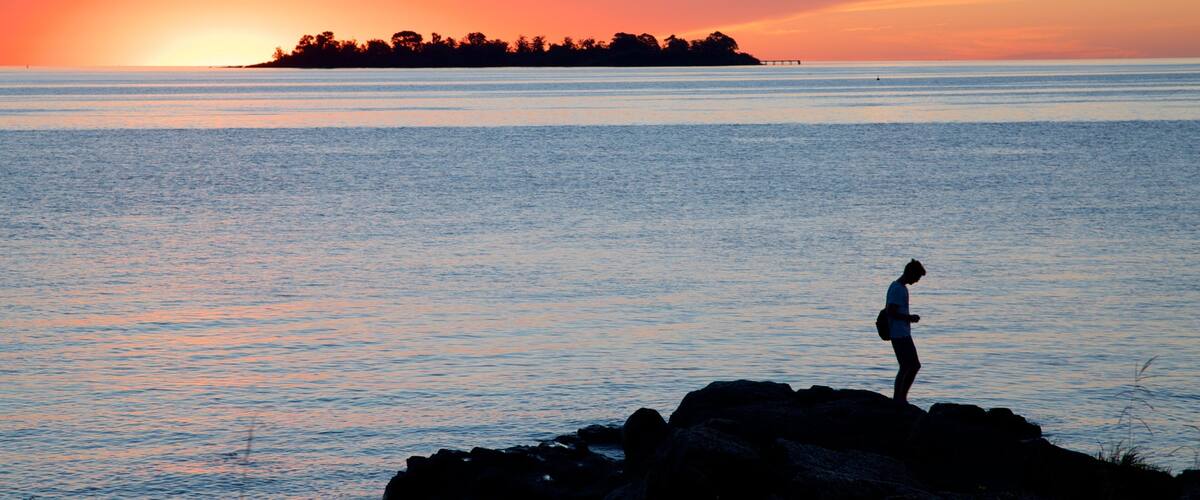 A person standing on rocks by the water during sunset.