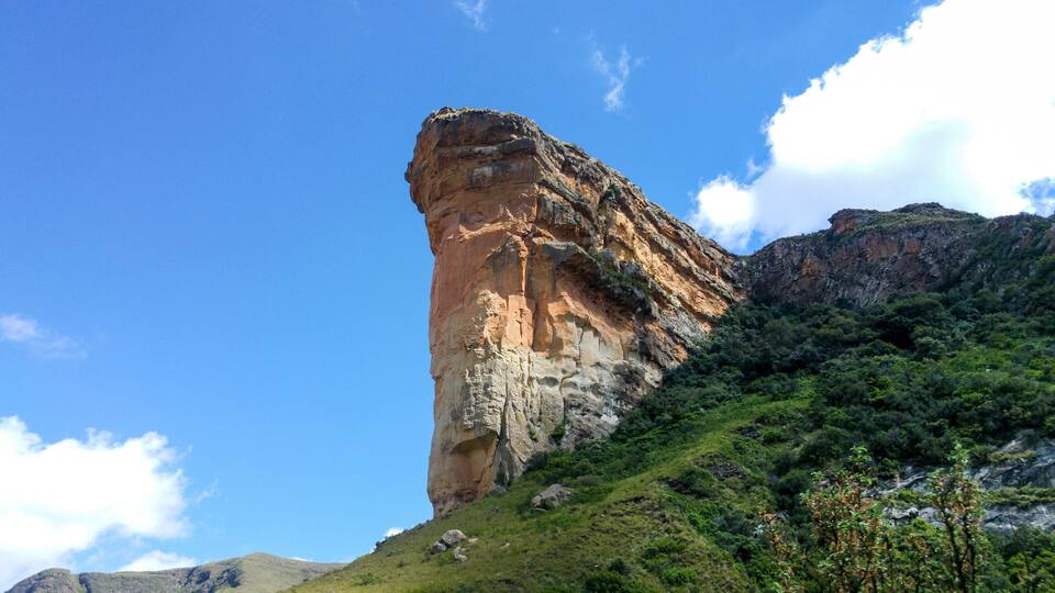A tall, narrow rock formation with a steep cliff face, surrounded by lush green vegetation and a clear blue sky.