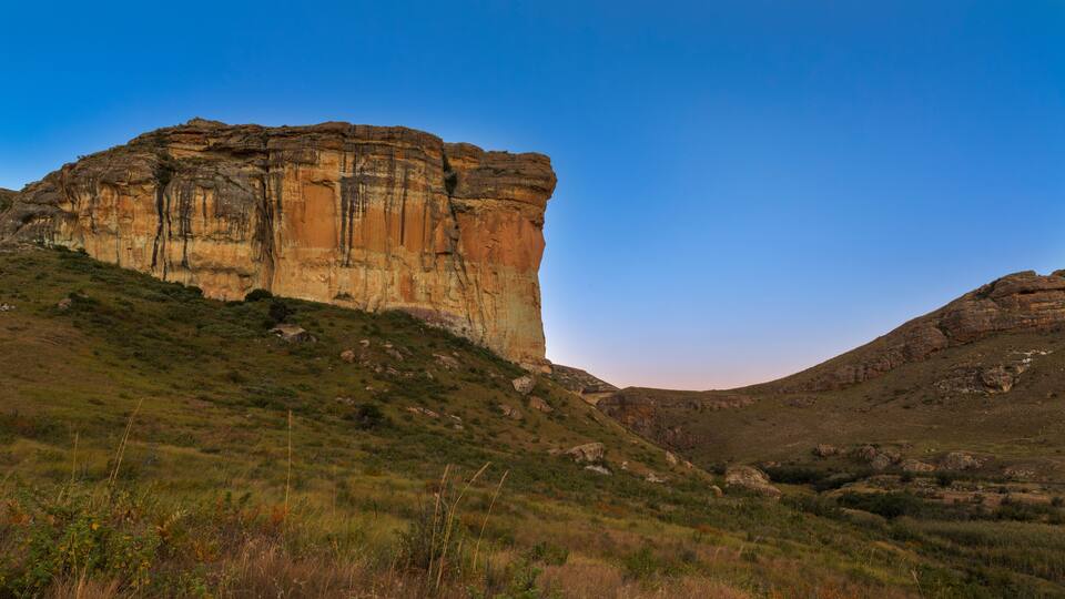 A large rock formation with layered sedimentary rock, surrounded by grassy hills and a clear blue sky.