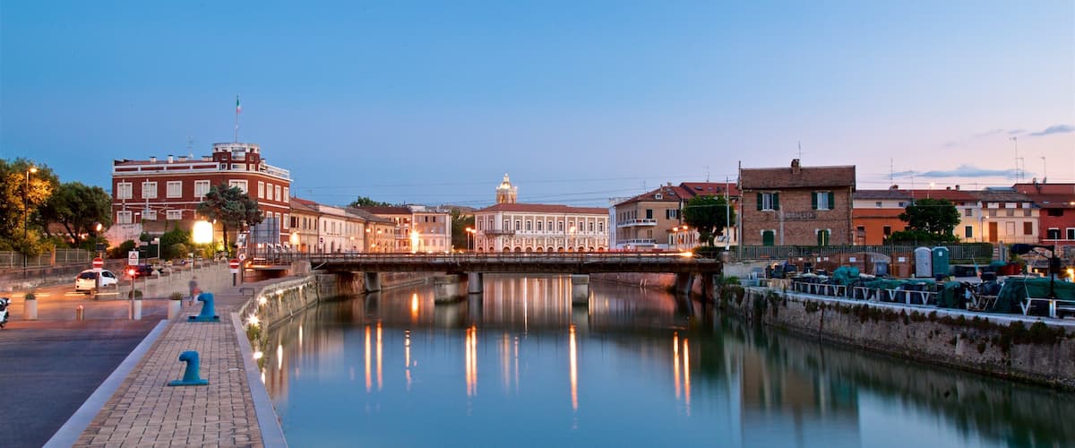 A calm river reflecting buildings and a bridge at dusk.