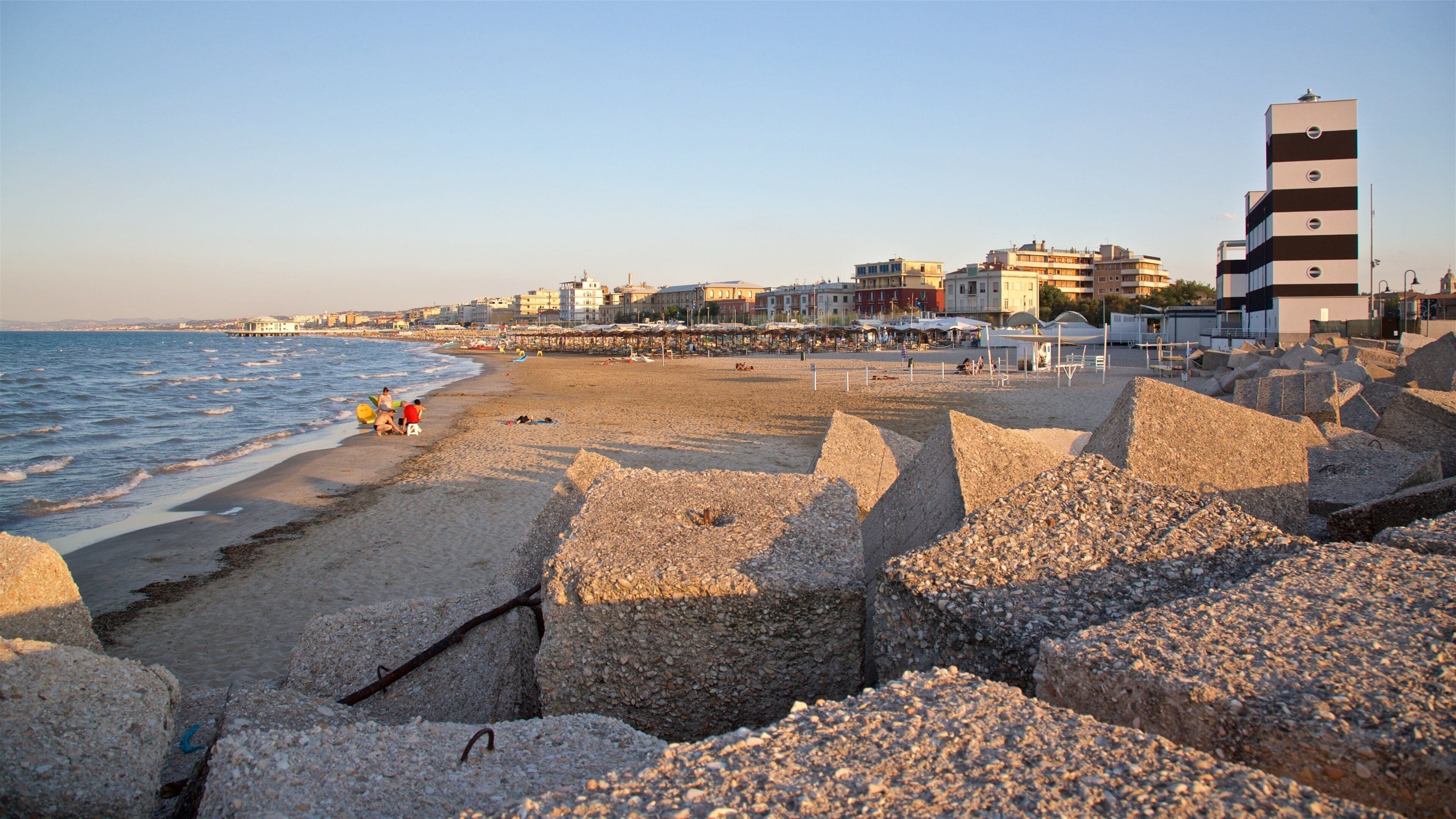 Una playa con rompeolas rocosas, un muelle y edificios al fondo.