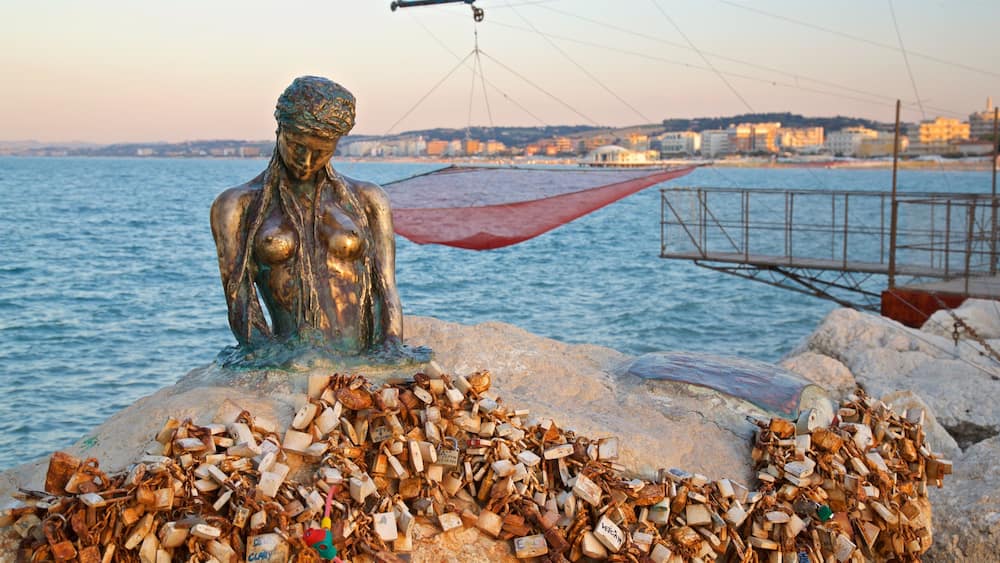 Eine Statue einer Frau mit einem roten Regenschirm an einer felsigen Küste am Meer, mit einer Stadtlandschaft im Hintergrund.