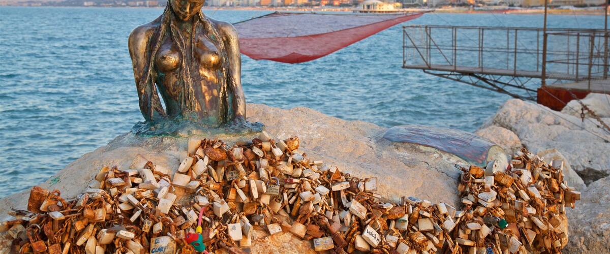 Een standbeeld van een vrouw met een rode paraplu op een rotsachtige kust langs de zee, met een stadsgezicht op de achtergrond.