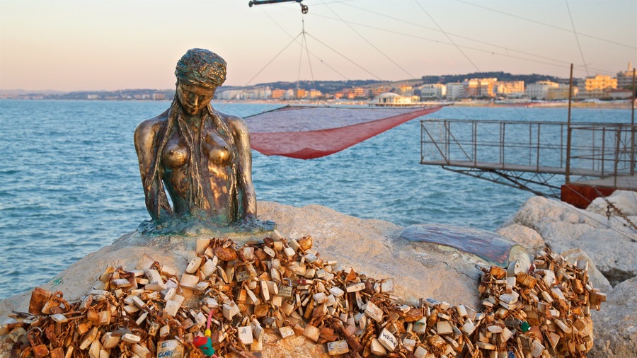 A statue of a woman with a red umbrella on a rocky shore by the sea, with a cityscape in the background.