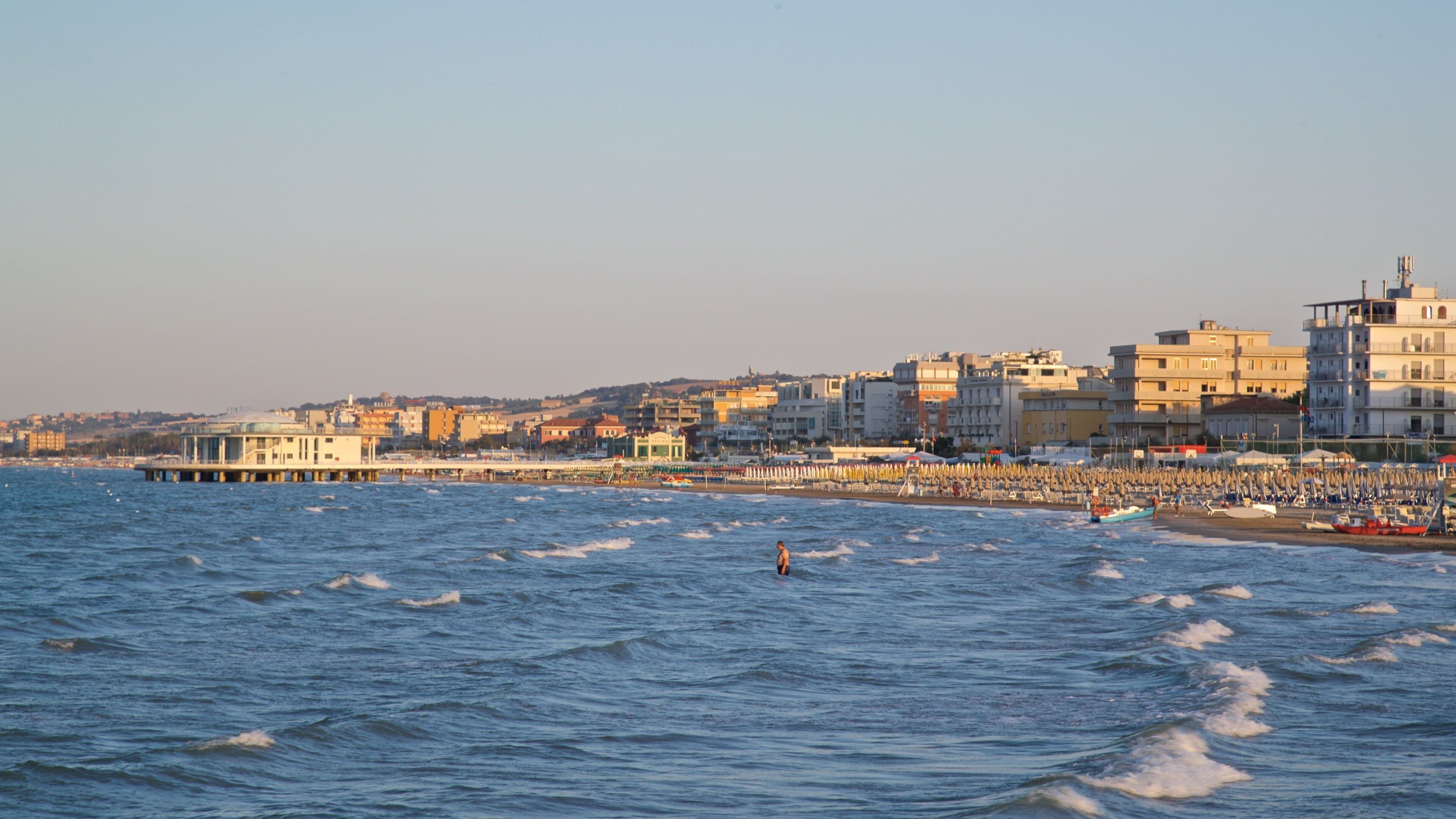 Un paisaje urbano costero con un muelle que se adentra en el mar, una playa llena de gente y edificios altos al fondo.