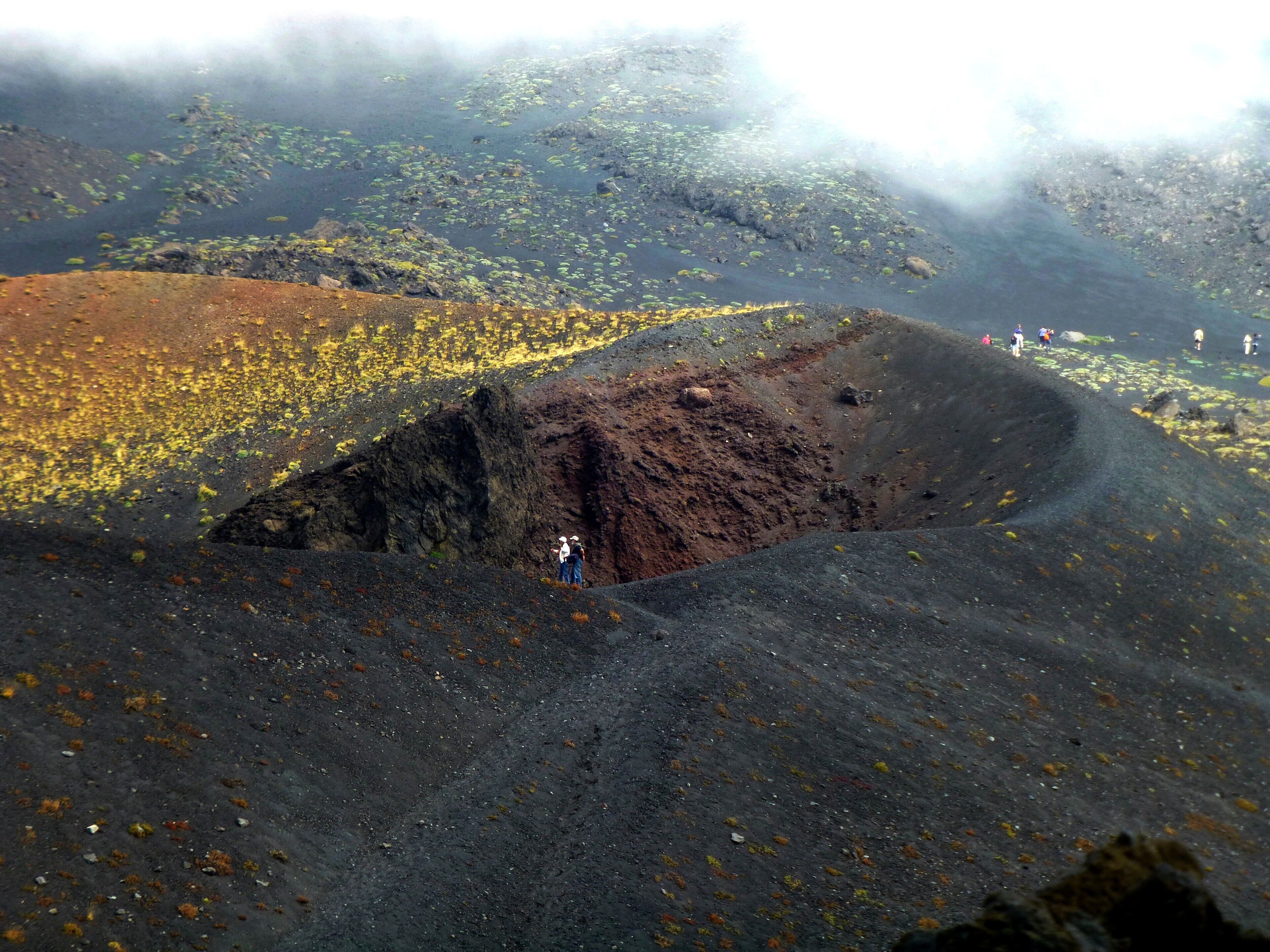 A volcanic crater with scattered vegetation and a few people standing on the edge.