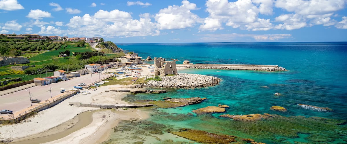 A coastal town with clear turquoise waters, a sandy beach, and a historic stone tower.