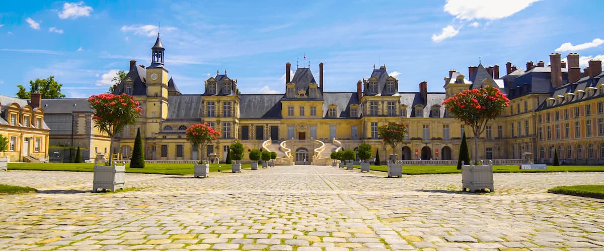 A historic courtyard with cobblestone paving, a grand building with multiple towers, and red flowering trees.