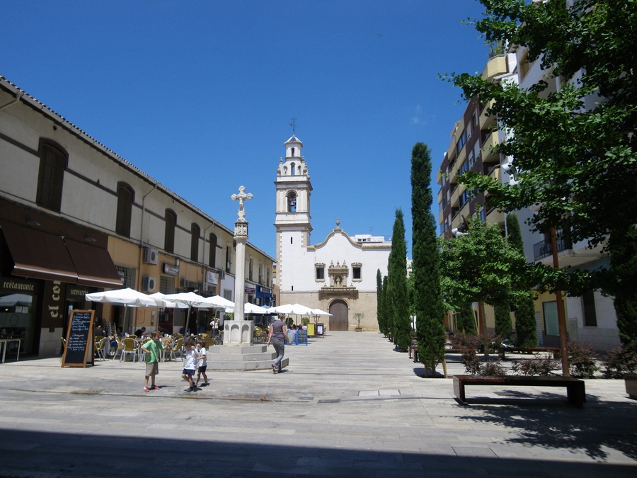 A plaza with a church tower, outdoor cafes, and trees.