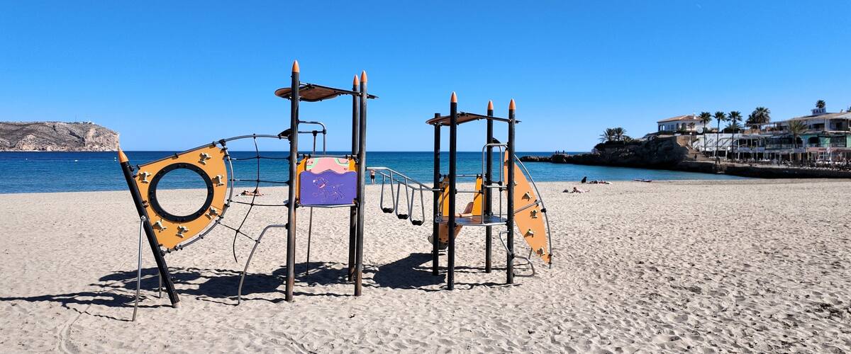 A beach playground with swings and climbing structures, a clear blue sky, and a rocky coastline in the background.