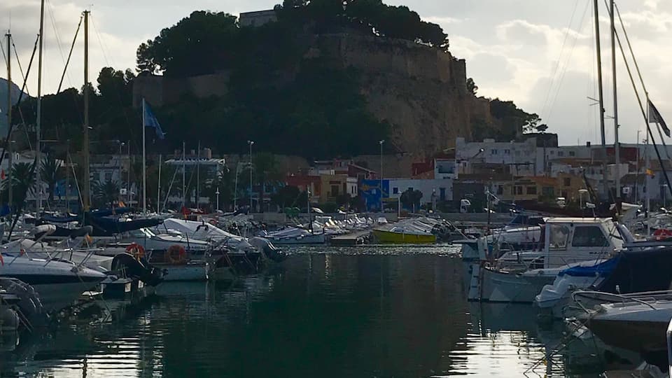 A marina with boats docked, a rocky hill with buildings in the background, and a cloudy sky.