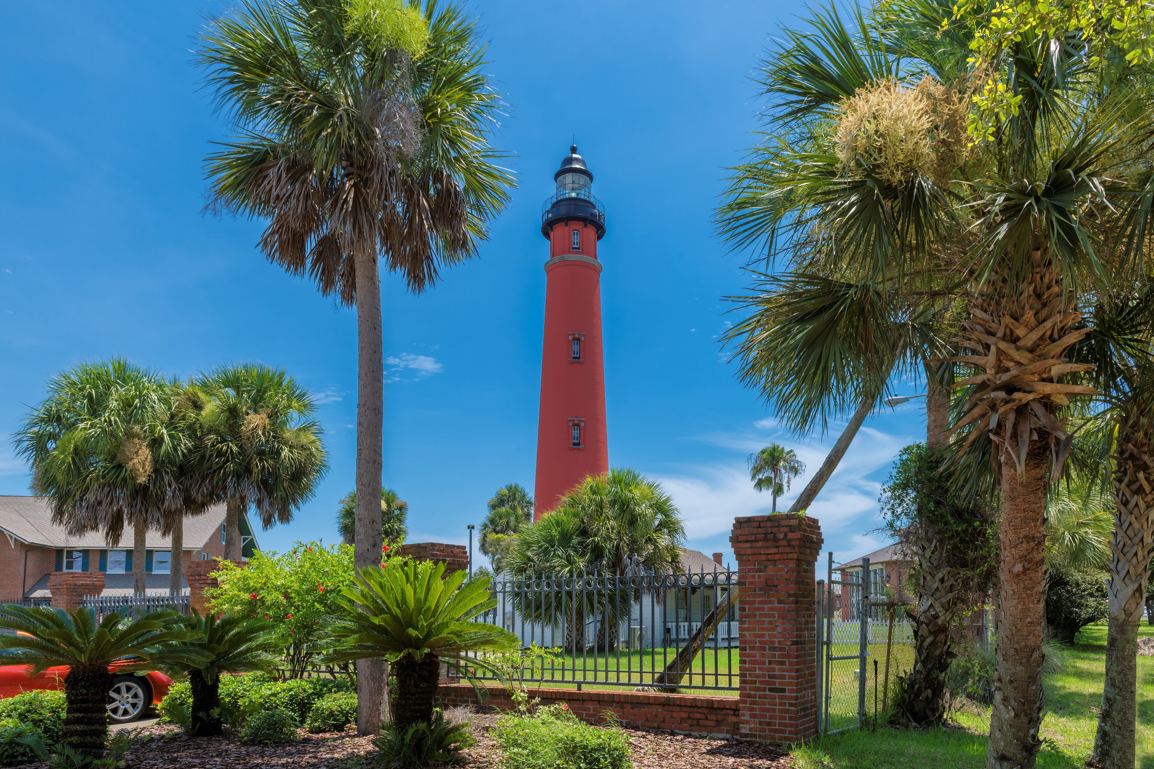 A red lighthouse surrounded by palm trees and a brick building.
