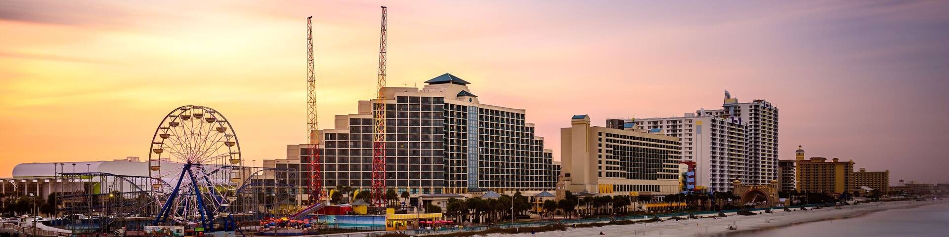 A coastal cityscape with a prominent Ferris wheel, high-rise buildings, and a beach at sunset.