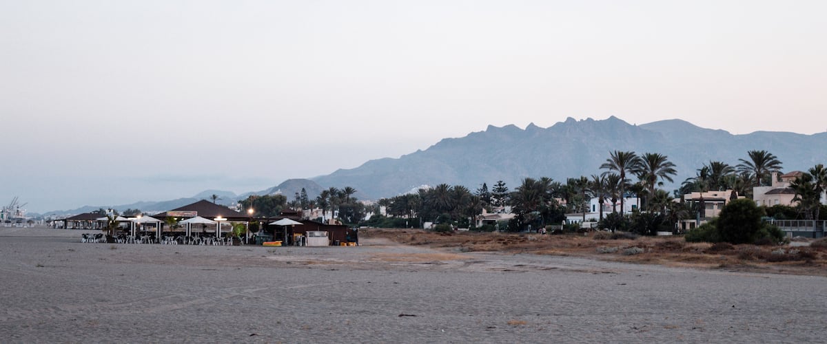 A beach with a row of beachside cafes, palm trees, and mountains in the background.