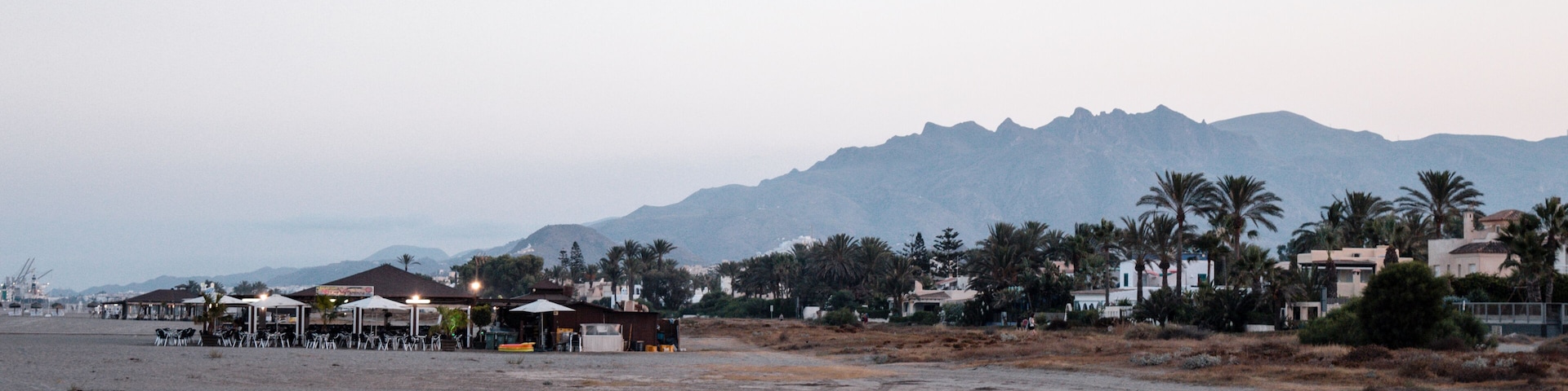 A beach with a row of beachside cafes, palm trees, and mountains in the background.