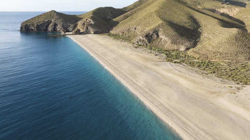 A coastal landscape with a sandy beach, clear blue waters, and green hills.