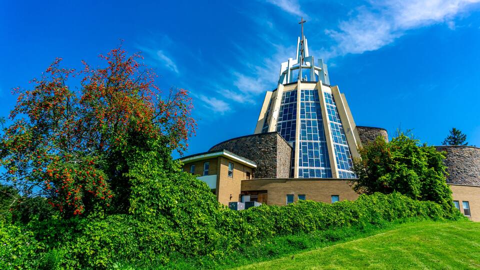 A church with a cross-topped tower, surrounded by lush greenery and a clear blue sky.