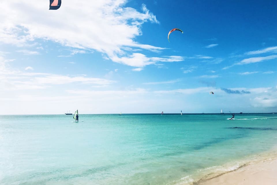 A beach with clear turquoise water, a sandy shore, and several people windsurfing.