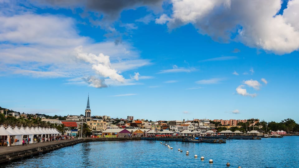 A coastal town with a prominent church spire, a pier with boats, and a clear blue sky.