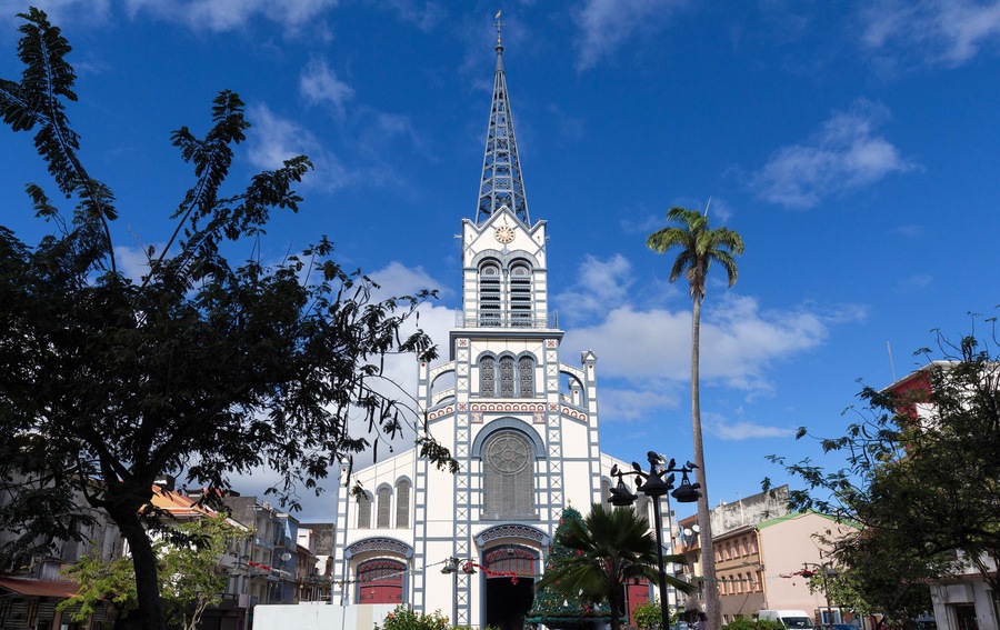Une église dotée d’un clocher élancé et d’une horloge, entourée d’arbres et de bâtiments.