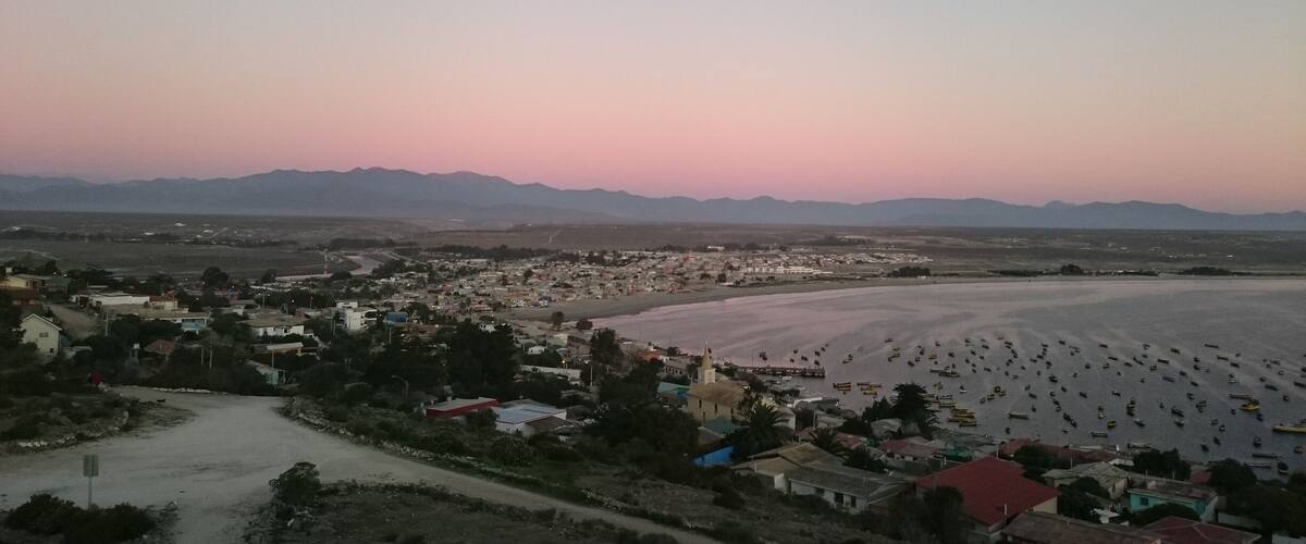 A coastal town with a beach, mountains in the background, and a clear sky.
