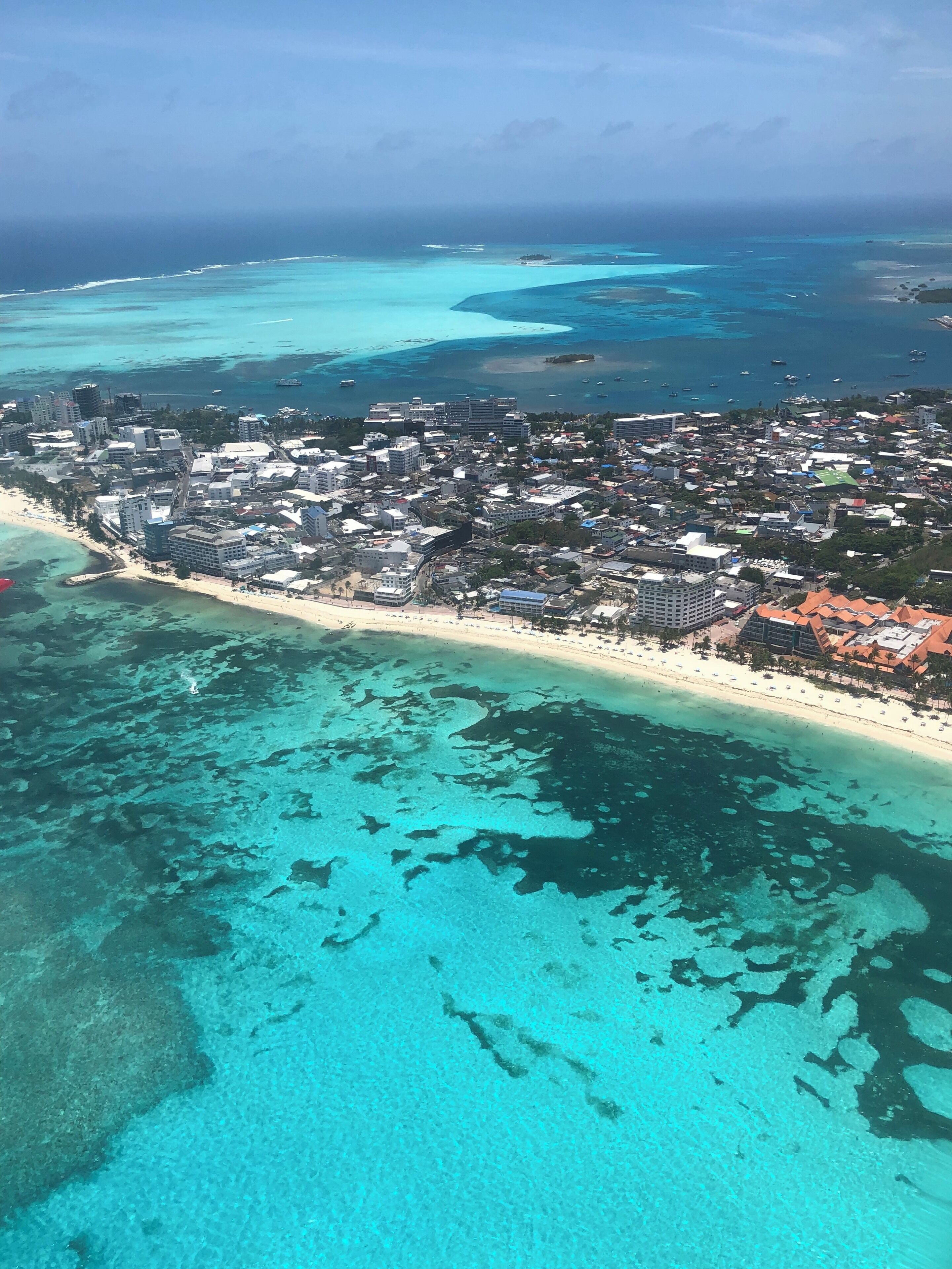 An aerial view of a coastal city with a beach, clear turquoise waters, and buildings.