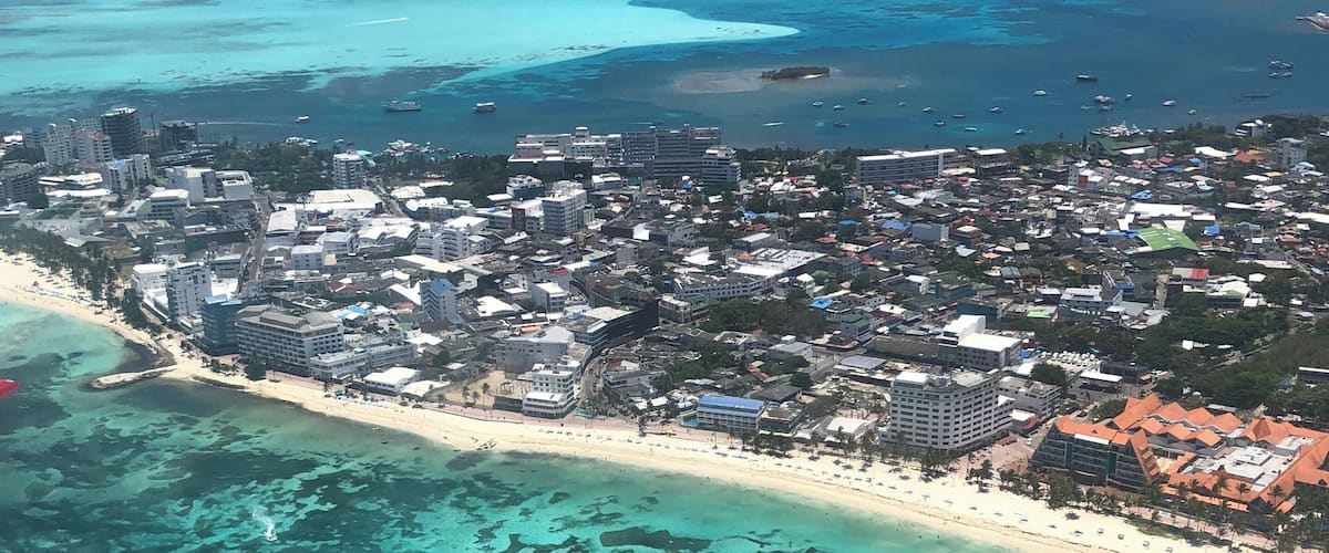 An aerial view of a coastal city with a beach, clear turquoise waters, and buildings.