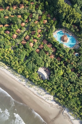 Une vue aérienne d’un complexe hôtelier comprenant plusieurs bâtiments de style bungalow, entourés d’une végétation tropicale luxuriante, avec une grande piscine et une plage.