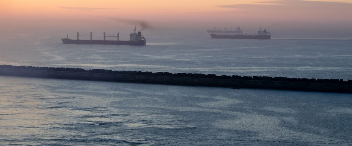 A large body of water with two cargo ships in the distance, a breakwater in the foreground, and a sunset sky.