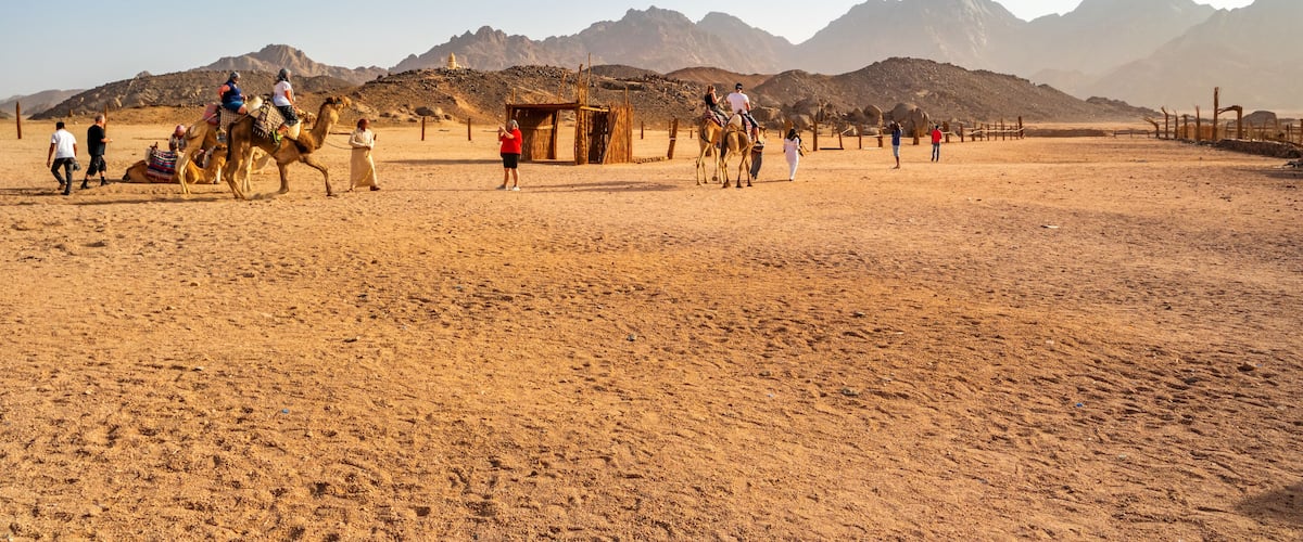 Paisaje desértico con gente montando camellos y una estructura de madera en el centro.