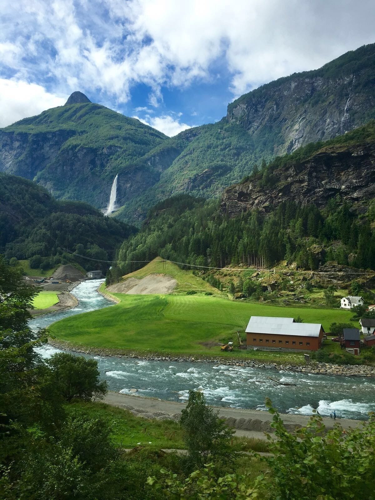A river flowing through a valley with a waterfall, surrounded by mountains and a small house.