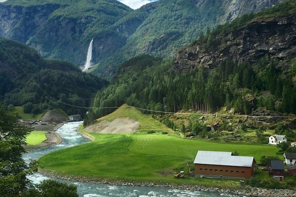 Ein Fluss fließt durch ein Tal mit einem Wasserfall, umgeben von Bergen und einem kleinen Haus.