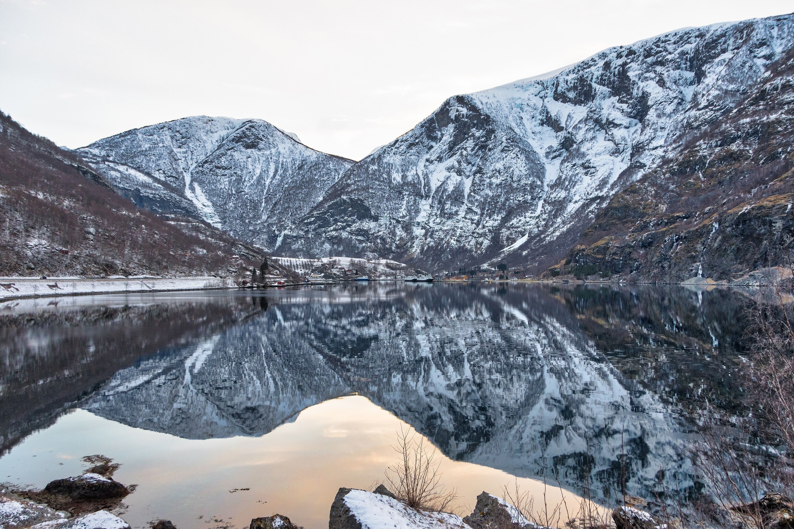 A snow-covered mountain range reflecting in a calm lake with rocky shores.