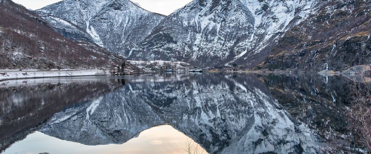 A snow-covered mountain range reflecting in a calm lake with rocky shores.