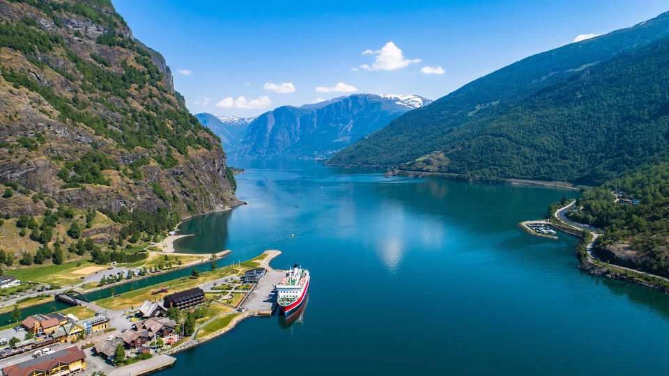 Un fiord amb una embarcació vermella i blanca amarrada a un moll, envoltat de muntanyes i un petit poble.