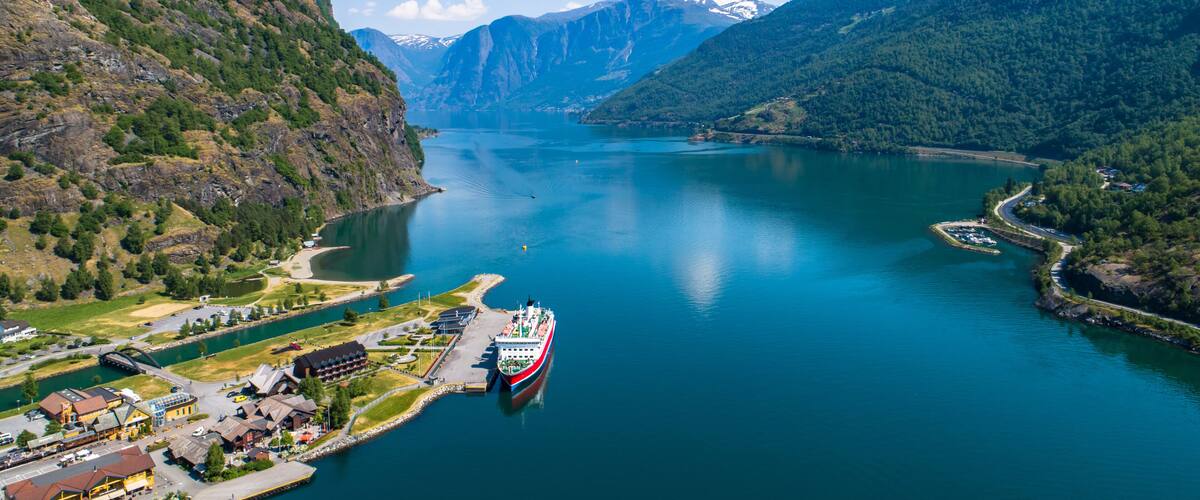 A fjord with a red and white boat docked at a pier, surrounded by mountains and a small town.