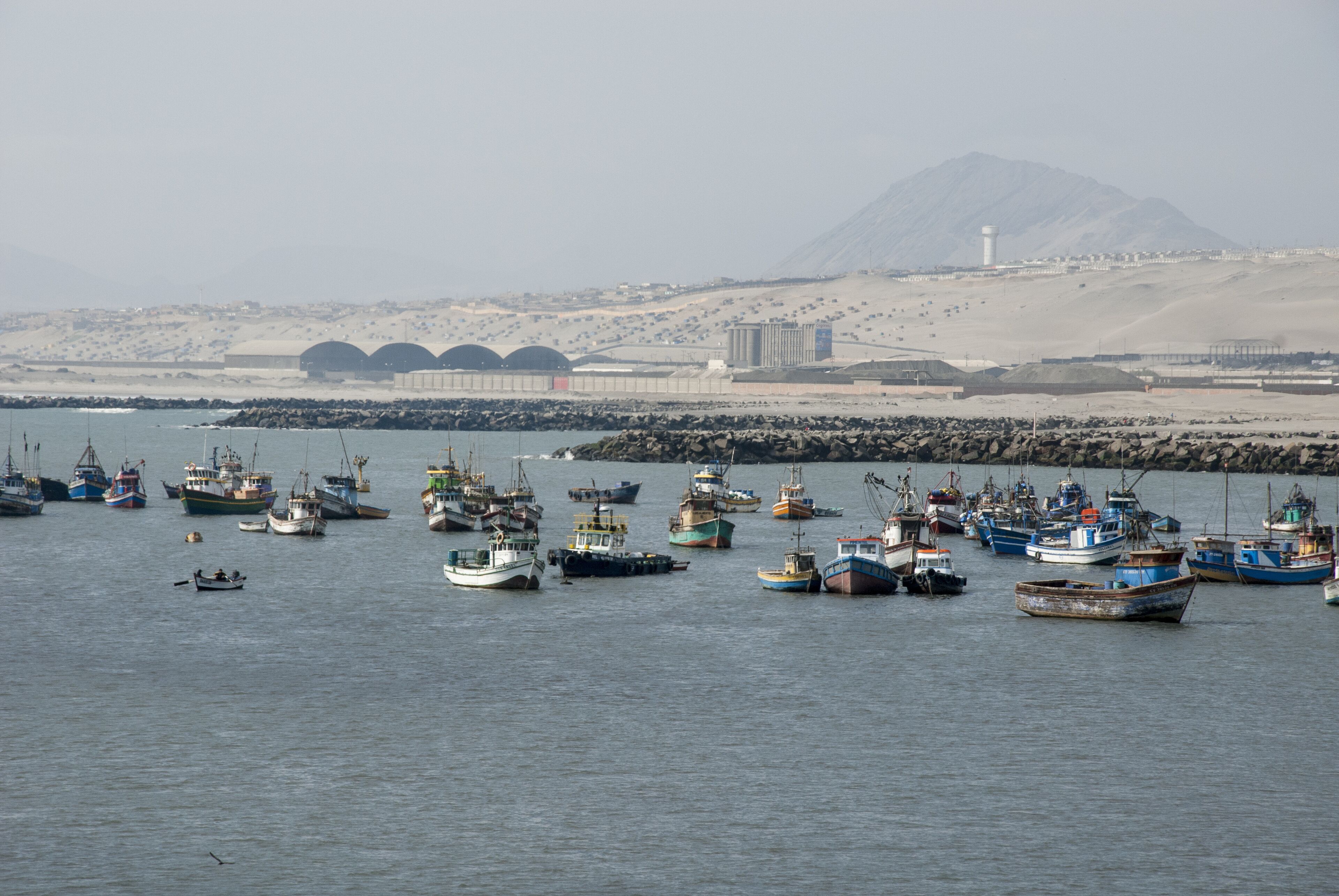 A harbor with numerous fishing boats, a lighthouse, and a mountainous landscape in the background.
