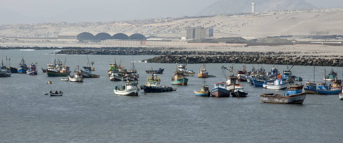 A harbor with numerous fishing boats, a lighthouse, and a mountainous landscape in the background.