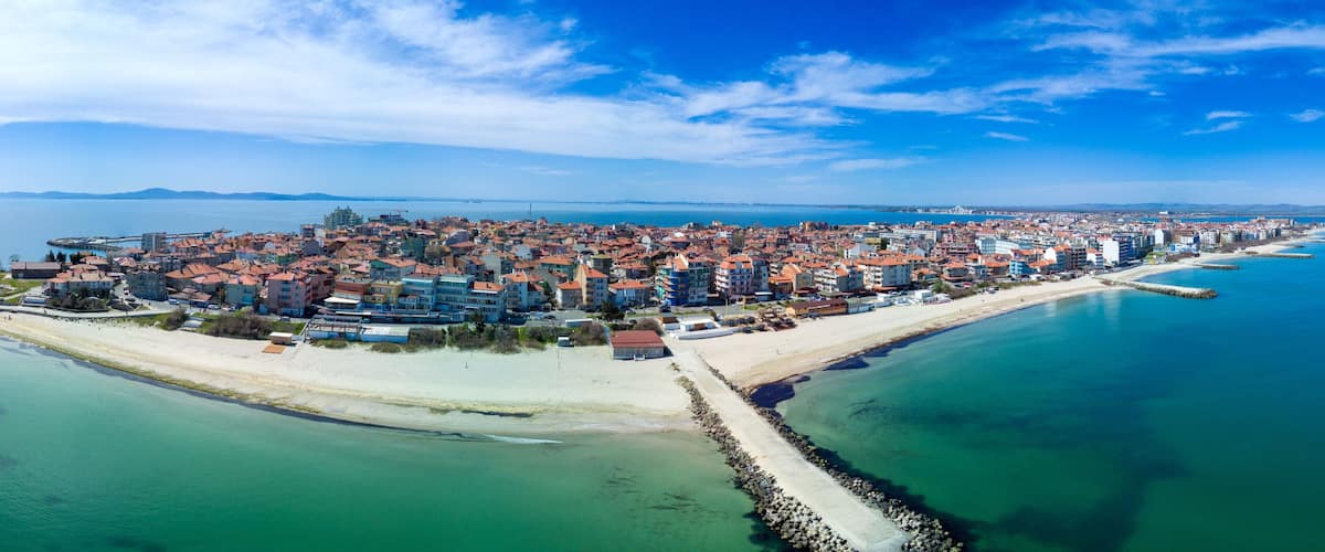 A coastal city with a prominent peninsula, a clear blue sea, and a distant mountain range.