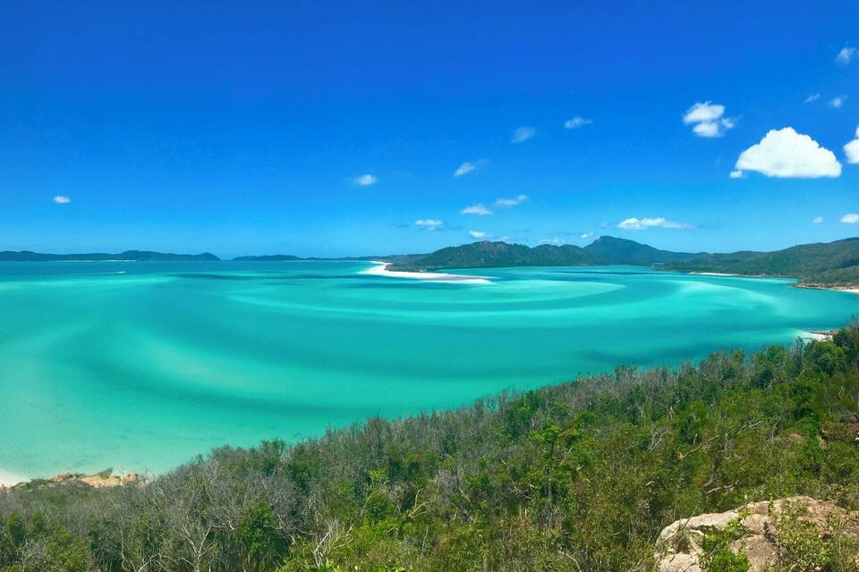 Una platja turquesa amb sorra blanca, envoltada per una forest verda i muntanyes al fons.