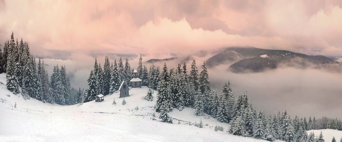 A snow-covered mountain landscape with a small cabin and dense evergreen trees.