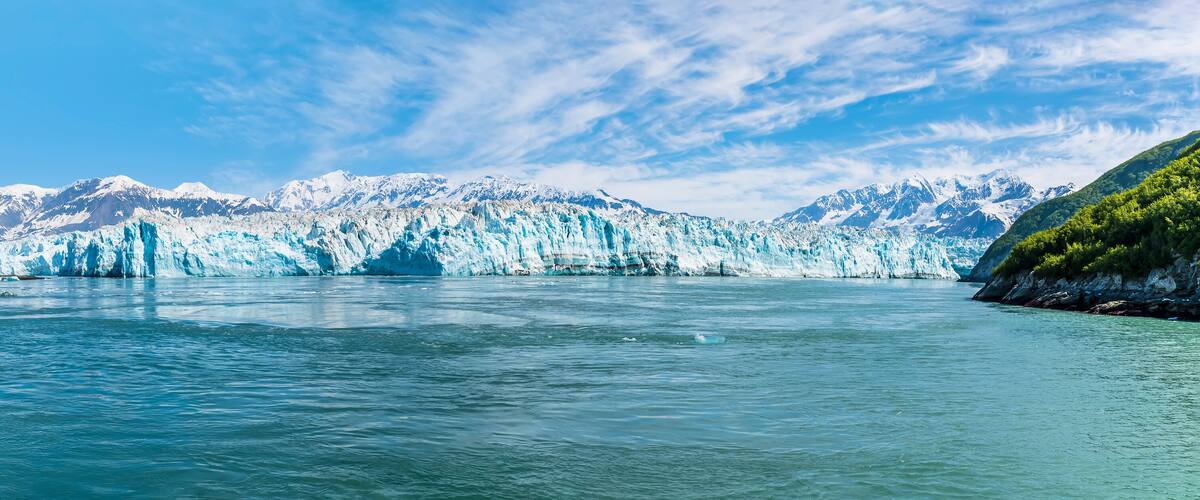A glacier with turquoise ice extending into a body of water, surrounded by snow-capped mountains under a clear sky.